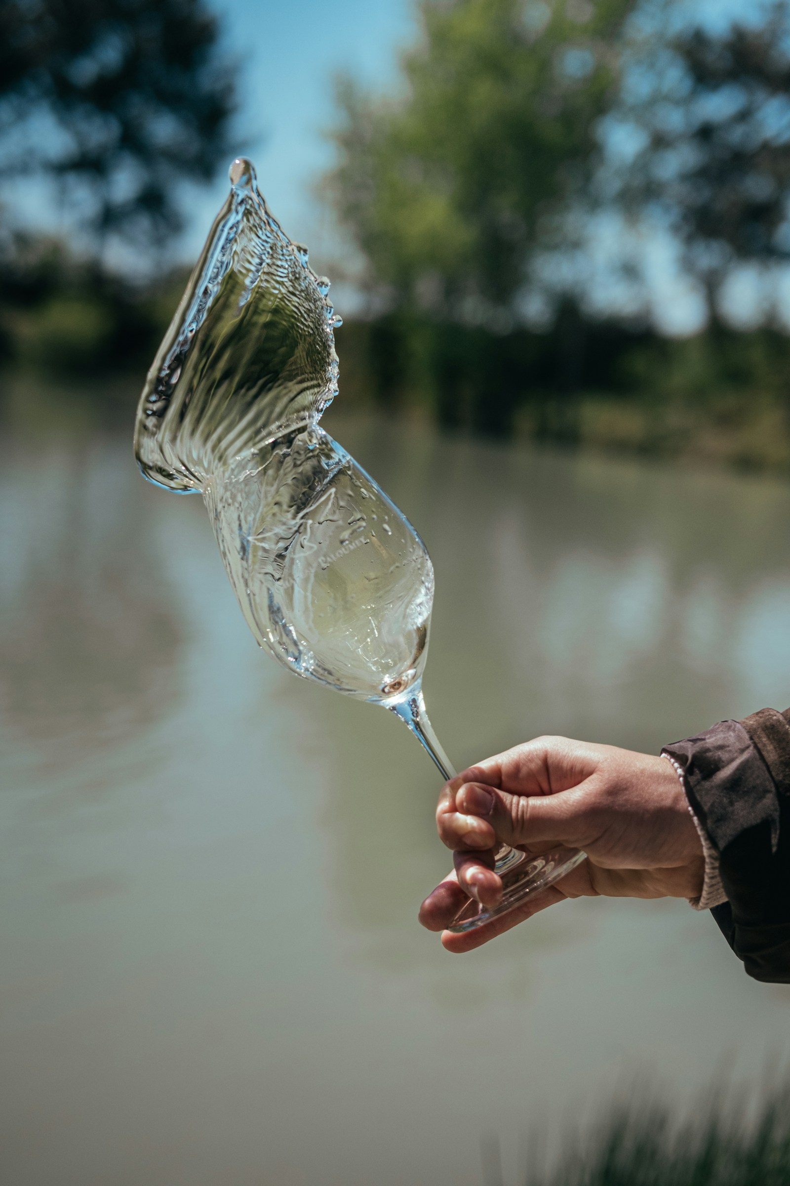 A glass of white wine held up beside a lake